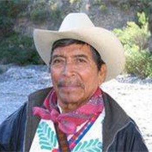 Pode incluir: Um homem usando um chap&eacute;u de cowboy claro, um len&ccedil;o estampado em vermelho e rosa e uma jaqueta preta. Ele tamb&eacute;m est&aacute; usando uma camisa branca com bordados coloridos. O fundo &eacute; uma cena externa desfocada.