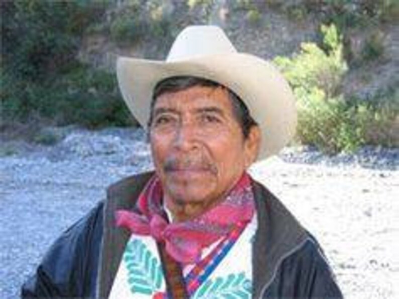 Pode incluir: Um homem usando um chap&eacute;u de cowboy claro, um len&ccedil;o estampado em vermelho e rosa e uma jaqueta preta. Ele tamb&eacute;m est&aacute; usando uma camisa branca com bordados coloridos. O fundo &eacute; uma cena externa desfocada.