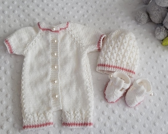 May include: White knitted baby outfit with pink trim and pearl buttons. The outfit includes a hat and booties.