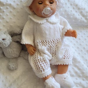 May include: A baby doll wearing a white knitted outfit, including a bonnet, romper, and booties. The romper features a scalloped collar and openwork detailing on the legs. The doll has a pacifier and is lying on a white, textured blanket.