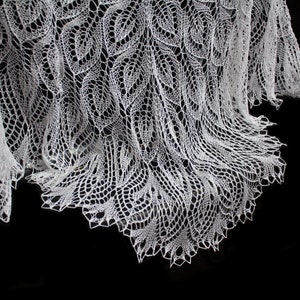 May include: A white, lacy shawl with a delicate leaf pattern. The intricate design is visible against a black background. The shawl's edges have a scalloped trim, adding to its elegant appearance.