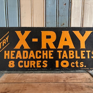 May include: Vintage black sign with orange text advertising "X-RAY HEADACHE TABLETS." The sign also states "8 CURES 10 cts." and "TRY" in a corner. The sign is displayed on a wooden surface.