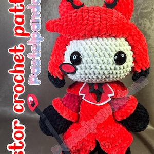 May include: A red and black crocheted plush toy of a character with horns, a white face, and a black bow tie. The toy is holding a black stick with a red flame on top. The toy is standing on a brown surface.