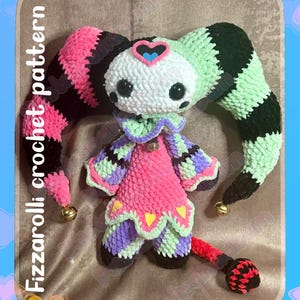 May include: A handmade crochet doll in a jester design. The doll features pink, black, green, purple, and white yarn. The doll has a heart-shaped face, large black eyes, and long, striped, floppy hat pieces. The text "Fizzarolli crochet pattern" and "Pastelpandabum" are visible.