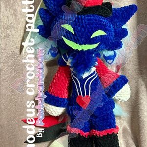 May include: A blue, red, and black crocheted plush toy with a top hat. The toy has a mischievous grin and a heart on its chest. The toy is labeled "Asmodeus crochet pattern by pastelpandabum".