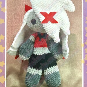 Peut inclure: Poup&eacute;e lapin en peluche faite &agrave; la main, avec un chapeau blanc et des accents rouges. Le lapin porte une tenue rouge et noire et a des bras et des jambes gris. La poup&eacute;e a de longues oreilles et un X rouge sur son chapeau. La poup&eacute;e est en mati&egrave;re douce et textur&eacute;e.