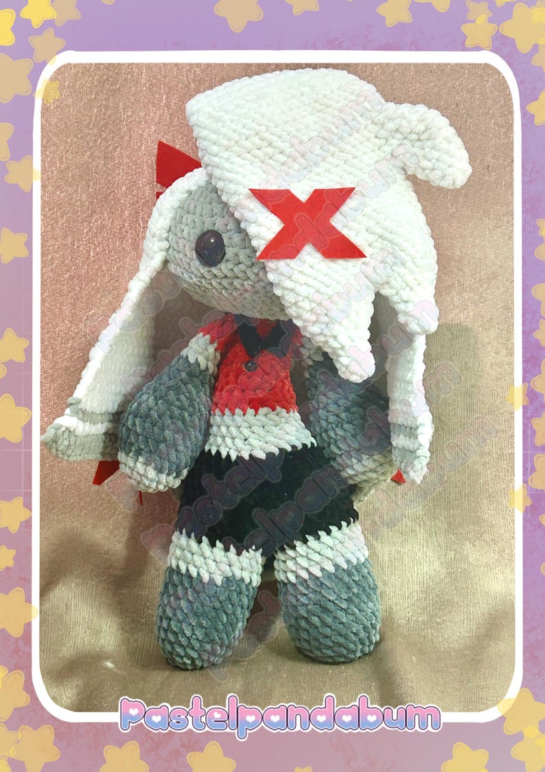 Peut inclure: Poup&eacute;e lapin en peluche faite &agrave; la main, avec un chapeau blanc et des accents rouges. Le lapin porte une tenue rouge et noire et a des bras et des jambes gris. La poup&eacute;e a de longues oreilles et un X rouge sur son chapeau. La poup&eacute;e est en mati&egrave;re douce et textur&eacute;e.