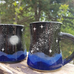 May include: Two ceramic mugs with a night sky design. One mug has a full moon and the other has stars. Both mugs have a blue mountain range at the bottom.