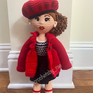 May include: A crocheted doll wearing a red beret, red jacket, black and white dress, and red shoes. The doll has brown hair and a crocheted design on the dress. The doll is standing on a wooden floor. The doll is labeled "@crochetsby_rae".