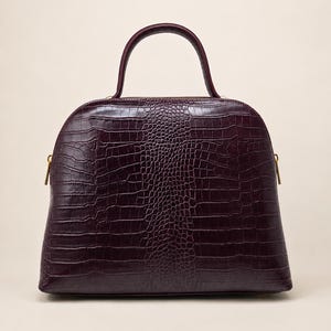 May include: A deep burgundy handbag with a structured dome shape and a top handle. The exterior features a textured crocodile-embossed pattern across the entire surface, complemented by gold-toned metal zipper pulls on the sides of the bag.