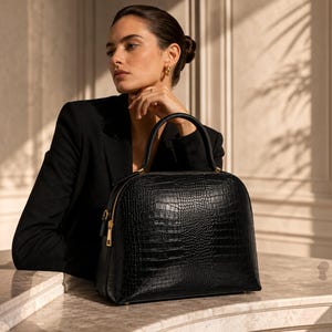 May include: A black handbag with a structured dome shape and a textured crocodile-embossed finish. The bag features a single top handle, a gold-toned zipper closure along the top, and gold-toned hardware accents on the sides and handle base.