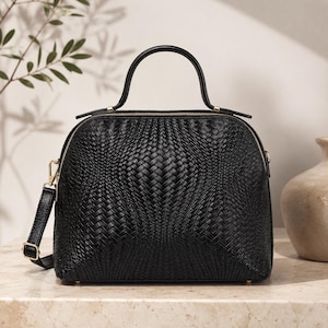 Peut inclure: Un sac à main noir doté d'une texture tressée sur le panneau avant, d'une poignée supérieure unique et d'une bandoulière amovible. Le sac présente une silhouette arrondie avec une fermeture éclair dorée et des détails en métal doré.