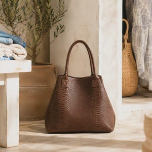 Peut inclure: Un sac cabas marron foncé avec un motif texturé en peau de serpent. Le sac présente deux anses verticales fixées sur le dessus, une base large et une silhouette structurée. La surface extérieure a un motif gaufré en écailles répétitives.