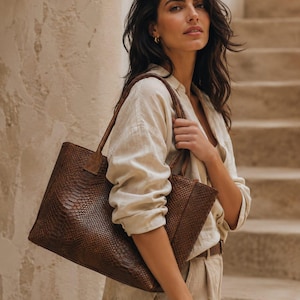 Peut inclure: Un sac fourre-tout en cuir marron avec un motif peau de serpent. Le sac a deux poignées et une forme rectangulaire. Le sac est porté par une personne vêtue d'une chemise et d'un pantalon en lin beige.