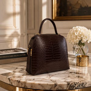 May include: A dark brown dome-shaped handbag with a textured crocodile-embossed pattern. The bag features a single top handle, gold-toned metal hardware, and a zippered closure. The structured design has a flat base and a smooth, polished finish.