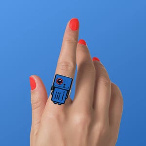 May include: A blue robot ring with a red eye and black outlines. The ring is worn on the index finger of a hand with red nail polish.