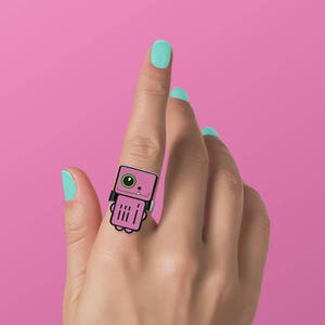 May include: A pink robot ring with black outlines and a green eye. The ring is worn on the index finger of a hand with light blue nail polish.