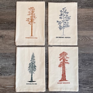 Puede incluir: Conjunto de cuatro toallas de cocina con ilustraciones de diferentes tipos de árboles. Cada toalla tiene un fondo marrón, crema o blanco con un contorno negro o marrón de un árbol. El texto "Ponderosa Pine", "Quaking Aspen", "Douglas Fir" y "Giant Sequoia" está impreso debajo de cada árbol.