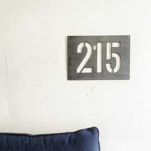 May include: A metal house number sign with the number 215 cut out of the metal. The sign is hanging on a white wall above a blue couch with a patterned pillow.