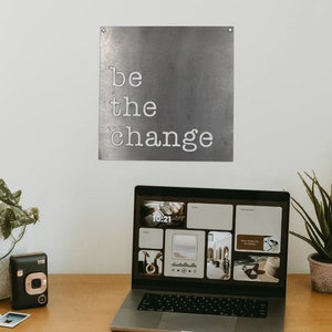 May include: A laptop computer with a motivational quote on the wall behind it. The quote reads "be the change". The laptop is open and has a screen with a collage of images. There is a potted plant and a camera on the desk.