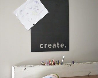 May include: A black metal wall-mounted chalkboard with the word "create" cut out in white. A piece of paper with a child's drawing is attached. A white distressed wooden organizer holds crayons and markers. A notebook is open on a wooden desk.