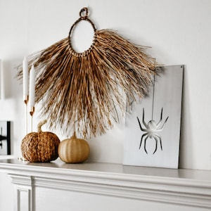 May include: A white painted wooden sign with a cutout of a black spider hanging from a string. The sign is on a white mantel with two woven pumpkins and a brown, dried grass wreath.
