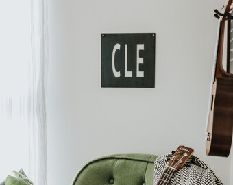 May include: A green upholstered armchair with a black and white patterned throw blanket and a wooden ukulele resting on top. A black metal sign with the word "CLE" in white letters is hanging on the wall behind the chair.