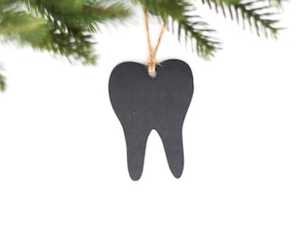 May include: A black tooth-shaped chalkboard ornament hanging from a branch of a green pine tree.