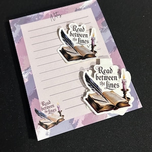 May include: A rectangular notepad with a purple and white watercolor border and the word Notes at the top. It features a graphic of an open book, a quill pen, and a lit candle with the text Read between the lines. Two matching stickers are placed on top.