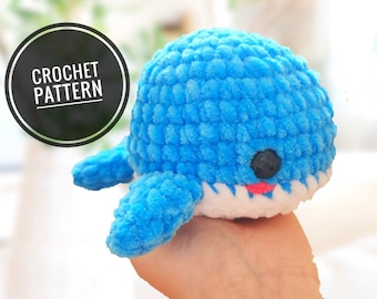 May include: A crocheted blue whale plush toy with white teeth and pink lips. The whale has black eyes and is made with a fluffy yarn.