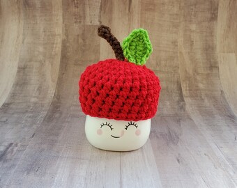 May include: A red crocheted apple hat with a green leaf and brown stem. The hat is sitting on a white mug with a smiling face.