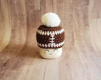 May include: A brown and white crocheted hat shaped like a football with a white pom pom on top. The hat is sitting on a white ceramic mug with a smiley face.