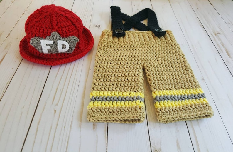 May include: A crocheted firefighter costume for a baby, featuring a red hat with a white "FD" patch and tan pants with yellow and gray stripes.