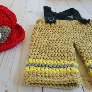 May include: A crocheted firefighter costume for a baby, featuring a red hat with a white "FD" patch and tan pants with yellow and gray stripes.