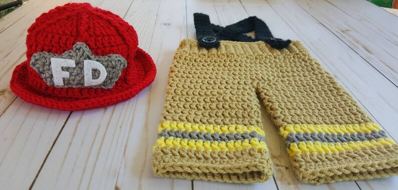 May include: A crocheted firefighter costume for a baby, featuring a red hat with a white "FD" patch and tan pants with yellow and gray stripes.