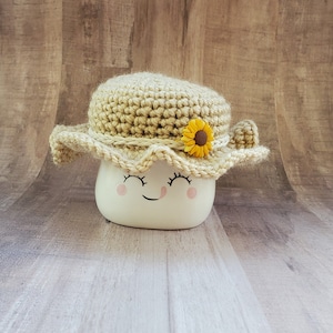 May include: A white ceramic mug with a happy face and pink cheeks, wearing a crocheted sun hat with a yellow sunflower accent.