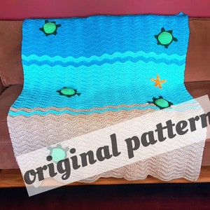 May include: A crocheted blanket with a beach scene featuring blue waves, tan sand, and green sea turtles. The blanket has the text "original pattern" on it.