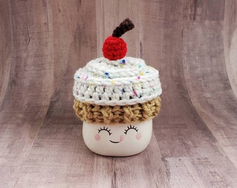 May include: A white ceramic mug with a smiling face, topped with a crocheted cupcake hat. The hat is white with colorful sprinkles and a red cherry on top.