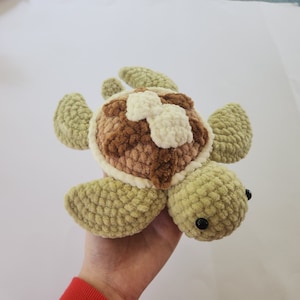 May include: A crocheted plush toy turtle with a brown and white shell and green body. The turtle has black eyes and a soft, fuzzy texture.