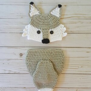 May include: A crocheted fox hat and matching swaddle blanket in a light green color with white accents. The hat has black eyes and a black nose.