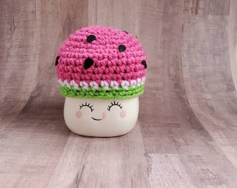 May include: A white ceramic mug with a smiling face, wearing a pink and green crocheted watermelon hat.