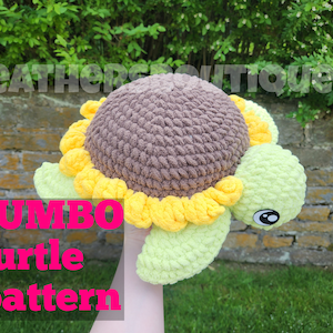 Puede incluir: Un juguete de tortuga de crochet con una concha marrón, pétalos de flores amarillos y patas verdes. El juguete se sostiene en una mano y el texto "JUMBO turtle pattern" es visible en primer plano.