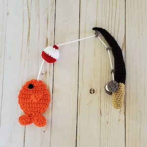 May include: A crocheted fishing rod with a red and white bobber and an orange fish. The fishing rod is made of black and beige yarn, and the bobber and fish are made of red, white, and orange yarn.