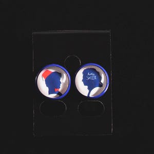 May include: A pair of blue and white round earrings with a silhouette of a person wearing a hat on one earring and the text "Little  Smith" on the other earring.