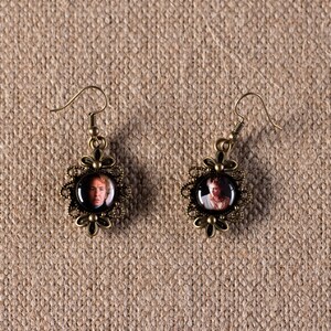 May include: A pair of gold-toned earrings with a round, black, framed image of a person's face. The earrings have a floral design.