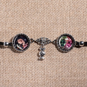 May include: A black leather bracelet with two silver round charms featuring colorful images. The charms are connected by a silver chain with a dangling silver charm.
