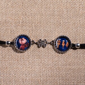 May include: A black leather bracelet with two silver round charms. One charm features a colorful image of a girl, and the other charm features a colorful image of three figures.