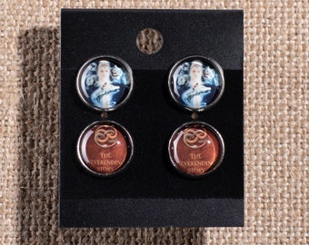 May include: Four round earrings with silver backing. Two earrings feature a blue and white image of a woman with a crown. The other two earrings feature a brown background with a gold infinity symbol and the text "The Everness Story".