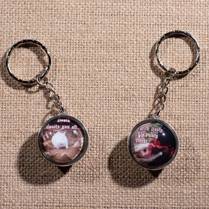 May include: Two round keychains with images and text. The first keychain has a white cat with the text "Jesus awaits you all." The second keychain has a cat wearing a Santa hat with the text "Wine Party, Ya'll Party,  Yeehaw!"