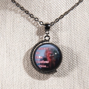 May include: A black metal necklace with a round pendant featuring an image of a bearded man and the text "Answer Me These Questions Three...". The pendant is encased in a decorative frame and hangs from a matching chain.
